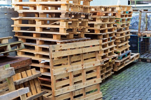 A large stack of wooden pallets arranged outdoors on a paved surface, with some stacked vertically and others horizontally, showing a natural unfinished wood texture with light brown and beige tones. The pallets have visible gaps between slats, with some displaying signs of minor weathering and staining. To the left, partial views of additional wooden items, possibly crates or pallets, are visible. In the background, there is a metal fence and a grey brick wall, with a slight indication of an urban environment, suggesting the location is near a commercial or industrial area in Byfleet. The ambient lighting is natural, highlighting the different textures and surfaces of the pallets. This scene aligns with waste management and rubbish removal services provided by Commercial Waste Byfleet, highlighting the handling of timber-based waste materials for disposal or recycling.