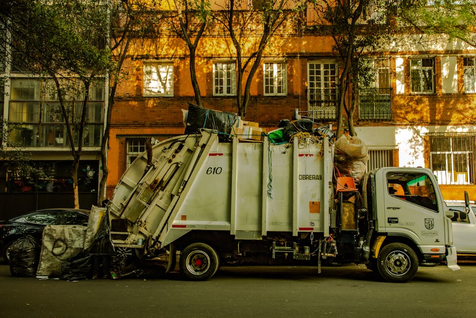A small waste collection truck from Obi Rea parked on a city street during daytime, partially surrounded by leafy trees and multi-storey residential buildings with large windows and balconies in the b