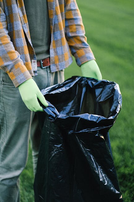 A person wearing a yellow and grey checkered shirt over a grey t-shirt, with olive green gloves, holds an open black rubbish bag by its edges outdoors on a grassy area. The bag appears to be made of p