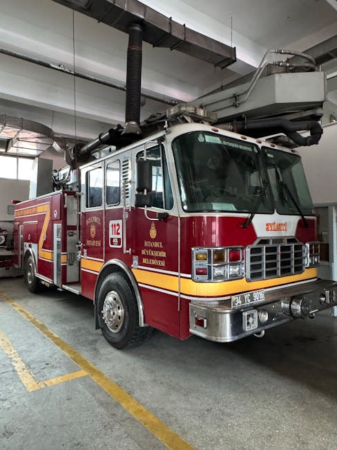 A red fire truck parked inside a garage or fire station, positioned on a concrete floor marked with yellow lines for parking bays. The vehicle features a white roof and chrome bumper, with multiple co